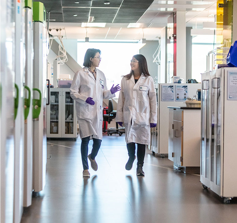 Two lab technicians in white lab coats having a conversation while walking in lab